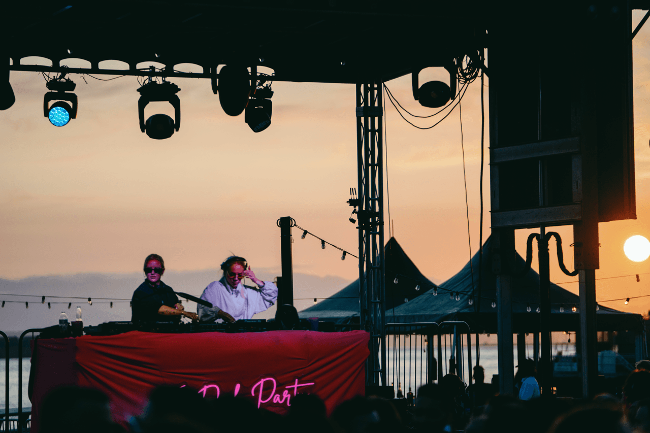 Two women DJing music to a crowd at sunset